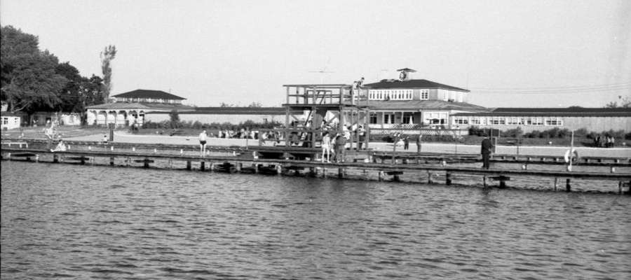  Plaża widziana od strony jeziora, zdjęcie wykonano prawdopodobnie z kajaka lub roweru wodnego. Na pierwszym planie skocznia i pomosty, dalej zabudowania z restauracją i przebieralniami. Lata 50. lub 60. ubiegłego stulecia